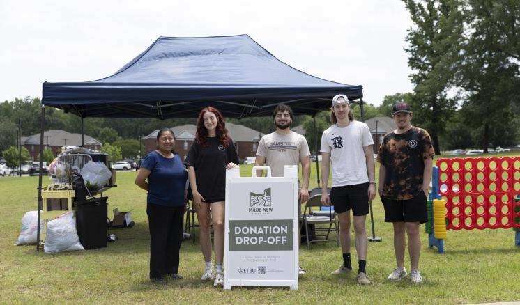Five people stand in front of a blue canopy tent at a donation drop-off event, with a sign reading "Donation Drop-Off" placed in the center.