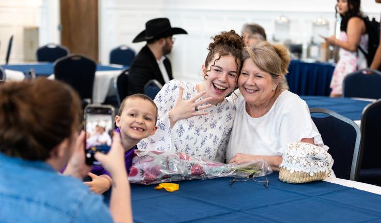 A woman taking a photo of 2 people and a child seated at a table with a blue tablecloth in a brightly lit room