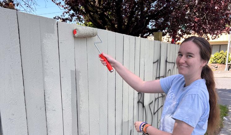 A woman using a paint roller to cover uop graffiti on a white fence