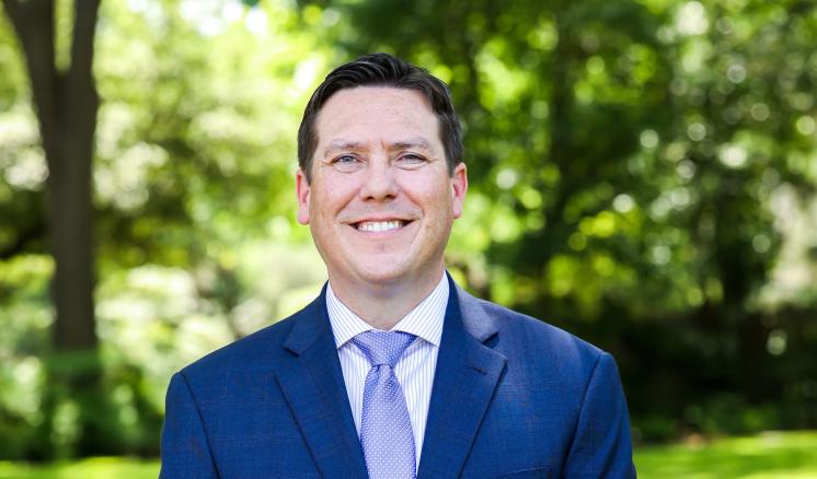 A man in formal clothing stood outside in front of trees on a walking path