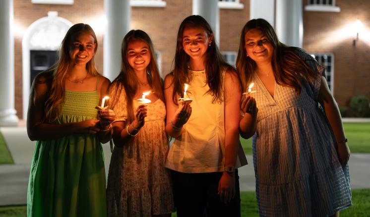 4 women standing together in front of a building outside with candles in their hands