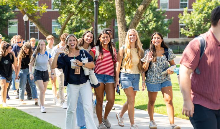 A group of students walking outside on a path smiling and waving
