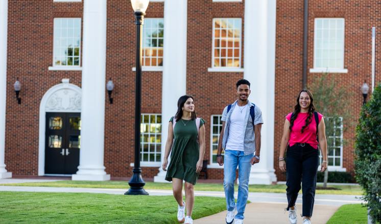 img_1.jpg Two females and one male ETBU student walking down a side walk in front of a red brick building with white columns in the background.