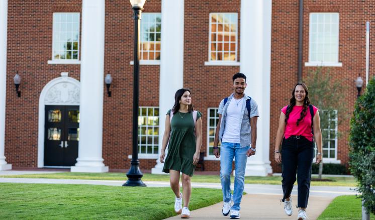 Two females and one male ETBU student walking down a side walk in front of a red brick building with white columns in the background.
