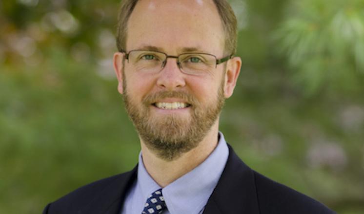 A man stood outside smiling in formal attire with a green backgrop of trees