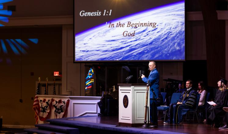 A speaker stood on a stage in formal attire with a projector screen showing Genesis 1:1
