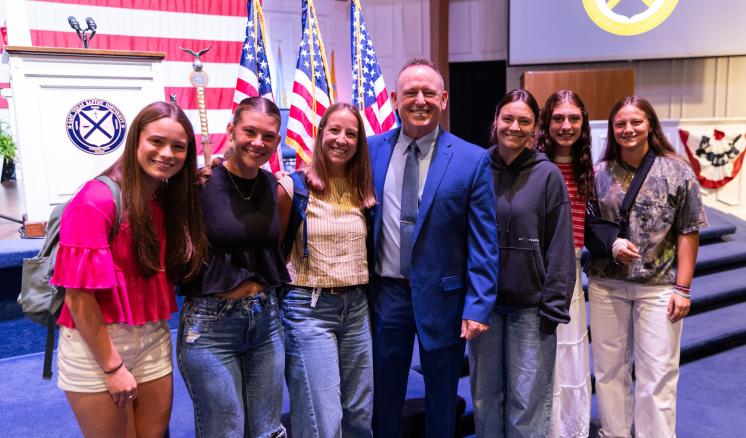 A group of people standing together smiling with the backdrop of American Flags on a stage