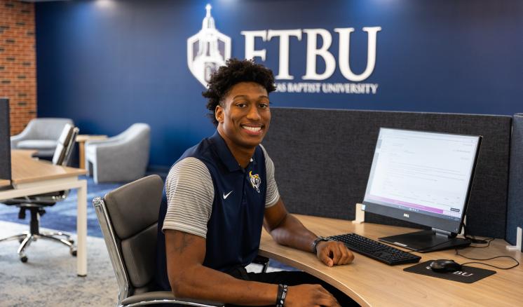 A man sitting at a table with a computer on it and the ETBU logo in the background