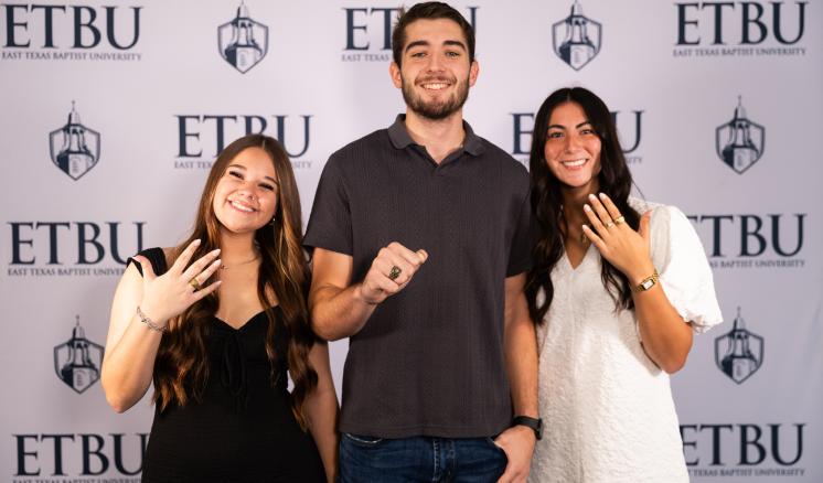 3 people standing in front of an ETBU backdrop with hands extended showing rings