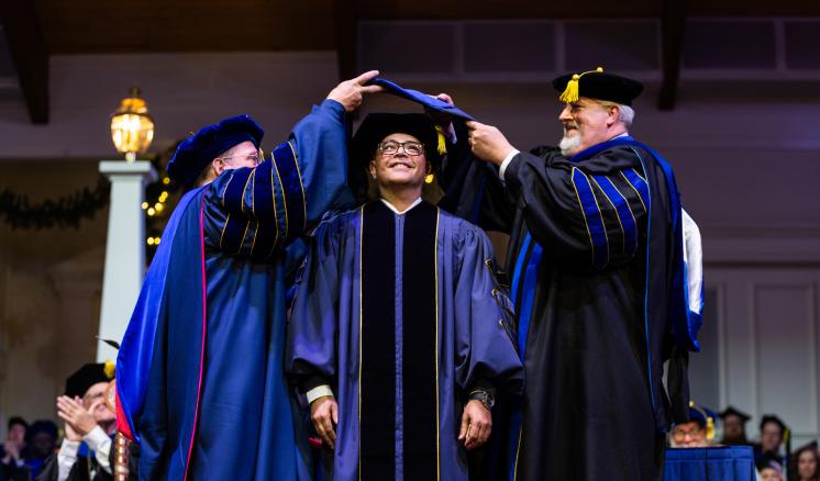 Three men in graduation regalia with the left and right men hooding a doctoral student in the middle.