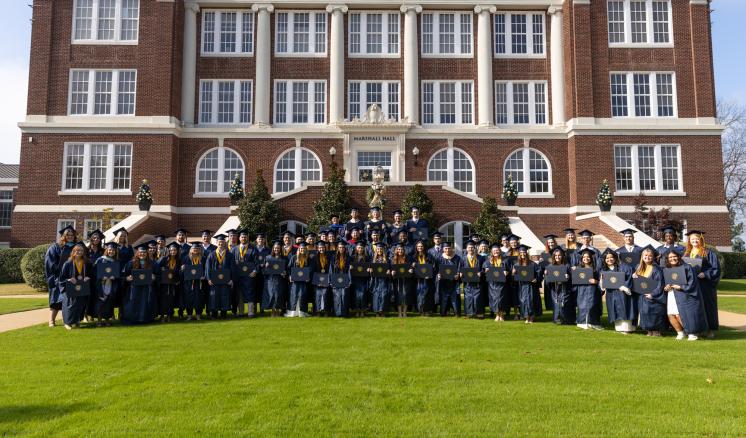 Group photo of graduates in navy regalia in front of Marshall Hall on a sunny day.