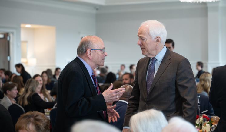 2 men standing talking together in a crowded room wearing professional attire