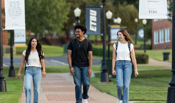 Three students walk down a campus sidewalk, smiling at each other.