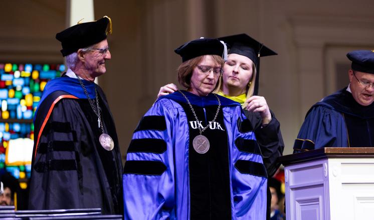 A woman putting a necklace with a large ETBU university seal around another womans neck wearing academic regalia