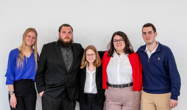 Five students stand side by side in professional attire and pose for a photo in front of a white wall.