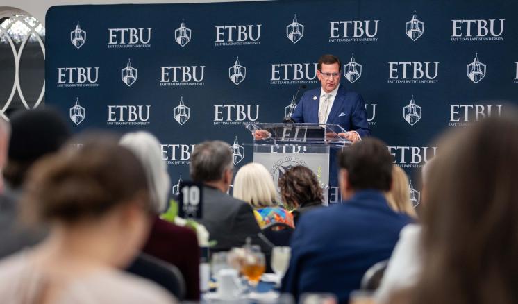 A group of people sitting and eating listening to a man speak behind a podium