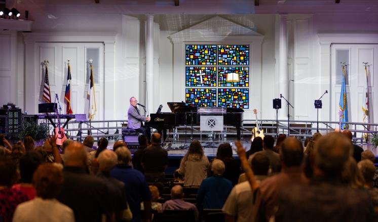 A pianist playing a grand piano in front of a colorful stained glass window depicting a cross and an open book, with an audience in the foreground.