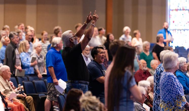 A diverse group of people standing and singing in an auditorium, with an older man raising his arms.