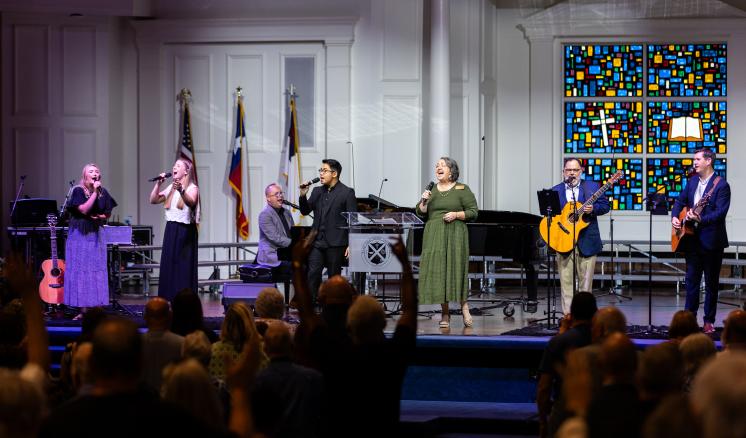 Performers singing and playing guitars on stage in a church setting with a stained-glass window in the background.