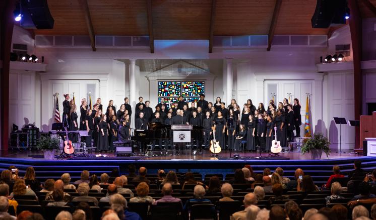 A choir performing on stage in a church-like setting, with a piano and stained glass window in the background.