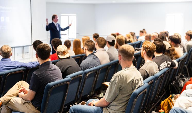 Rows of students are seated in blue chairs listening to a speaker who is standing in front of a projector screen.