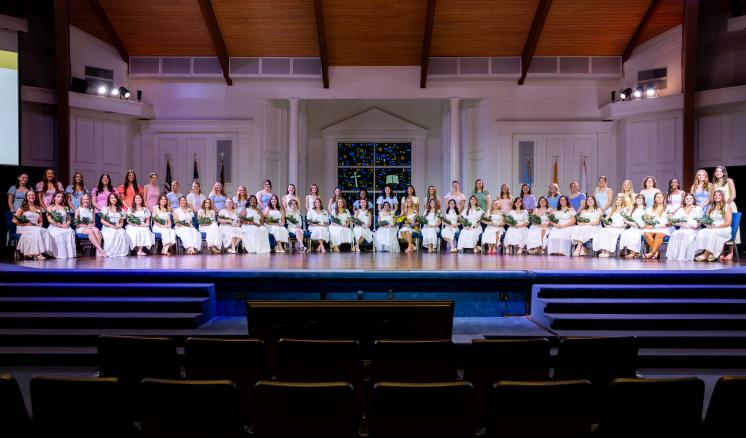 A large group of women smile on a stage for a photo. The wide shot features one row of women in white dresses seated with another row of women standing behind them.
