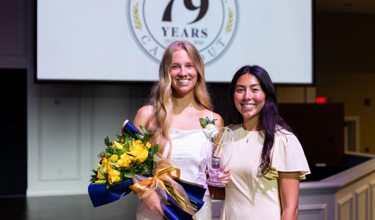 Two girls stand in white dresses. One is holding a bouquet of flowers and there is an event slide shown on a projector screen behind them.