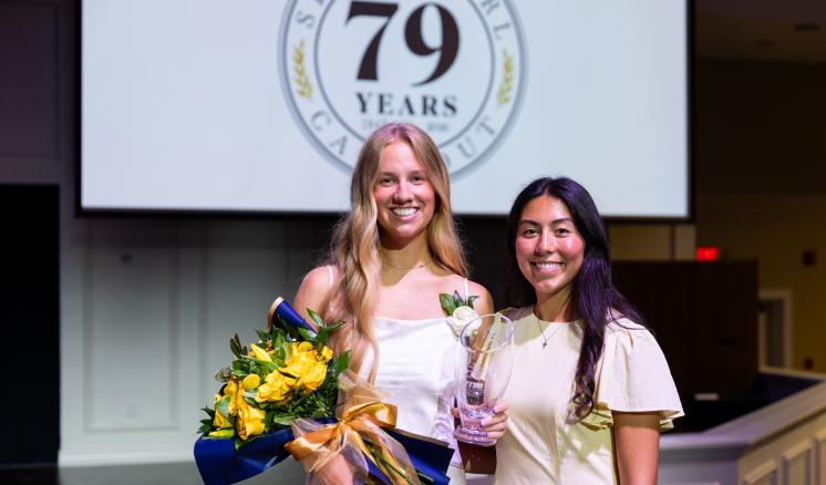 Two girls stand in white dresses. One is holding a bouquet of flowers and there is an event slide shown on a projector screen behind them.