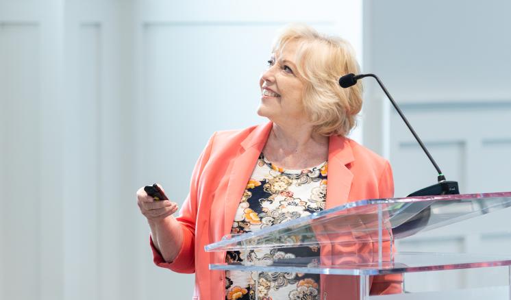 A woman in a pastel blazer stands behind a podium, she smiles up and points a remote to a projector