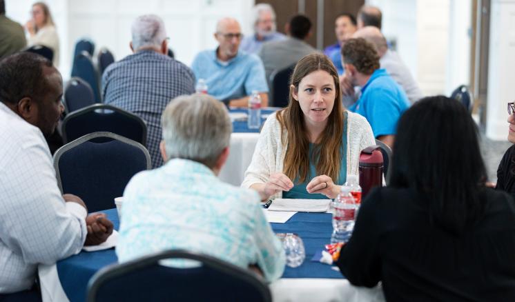 A woman sits at a table with a group of people. She is talking to the group.