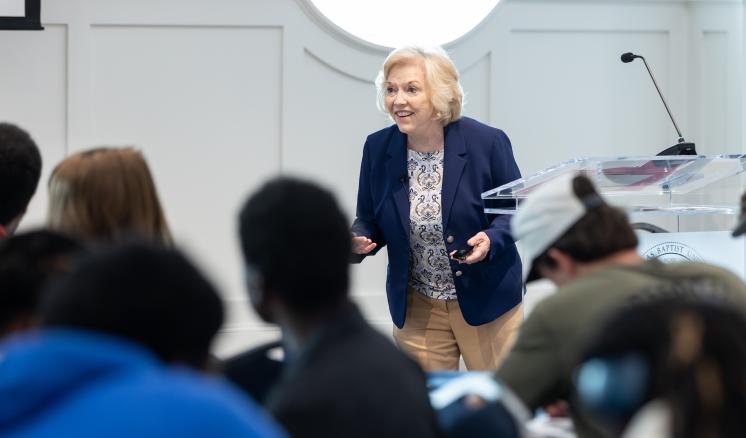 A woman in a blue blazer stands behind a podium talking to an audience of people in a brightly lit room