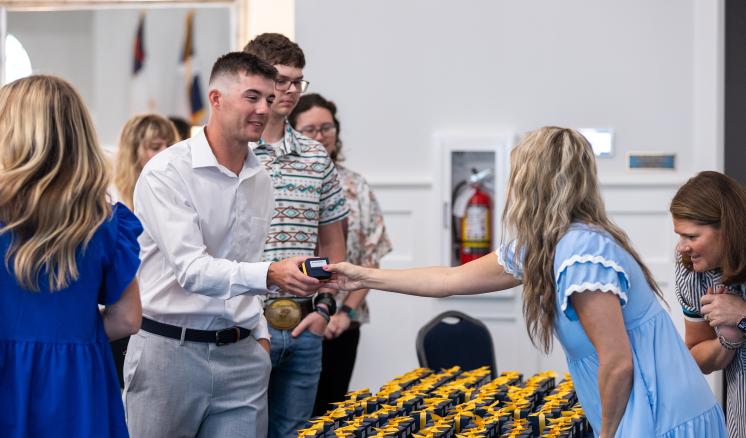A student smiles as he is handed his class ring. A table with several ring boxes is shown pictured in front of him.