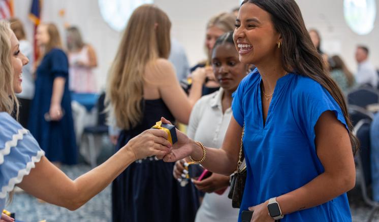 A student in a blue blouse smiles as she is handed her class ring in a box.