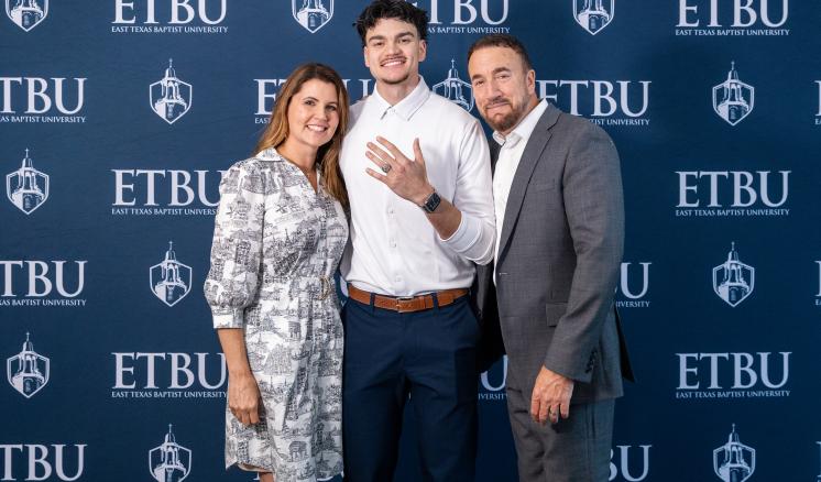 One student and his parents stand in front of an ETBU-branded backdrop. The student shows his class ring