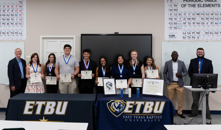Students and faculty in a classroom stand behind a table with ETBU branded tablecloths. The students are smiling and holding awards.