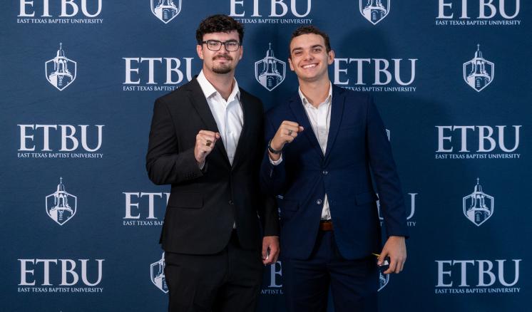 Two students stand in front of an ETBU-branded backdrop showing their class rings