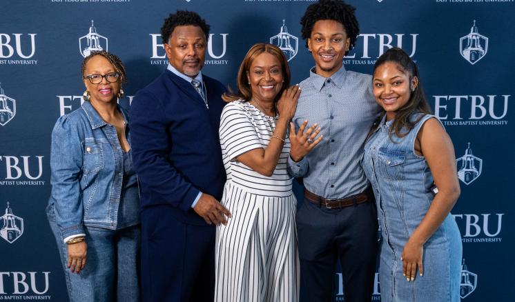 One student and his family, four other individuals, stand in front of an ETBU-branded backdrop. The student is showing his class ring.
