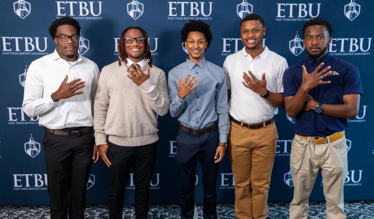 Five students stand in front of an ETBU-branded backdrop showing their class rings
