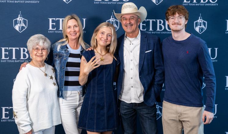 A student stands in front of an ETBU-branded backdrop with her family of four other people. She is showing her class ring.