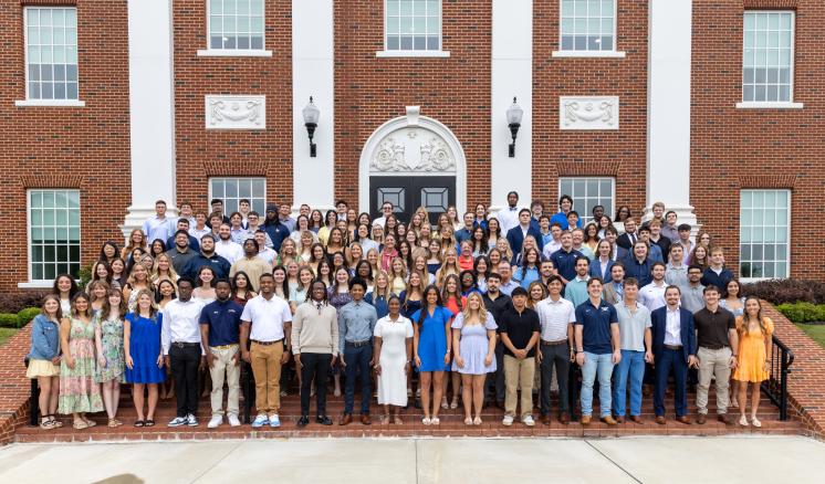A large group of more than 100 students stands on an exterior staircase showing their class rings