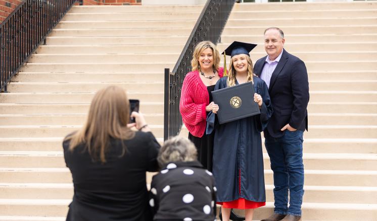 Group photo of a graduate in navy regalia with her family on the steps of a red brick building