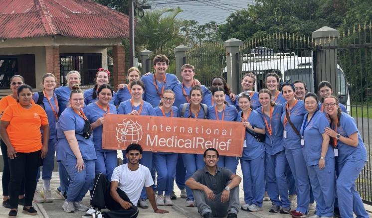 A group of students in blue scrubs stand with a International Medical Relief orange sign in front of a scenic backdrop.