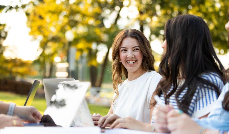 Photograph of young adults studying outdoors with laptops and notebooks, capturing a moment of smiling and engaged conversation. Warm sunlight and autumn foliage create a relaxed, collaborative learning environment.