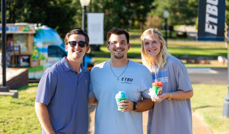 Photograph of three young adults standing outdoors on a sunny day, smiling and holding colorful shaved ice treats. Background includes a shaded park area, a food cart, and a vertical banner with "ETBU" text, indicating a casual social or campus event.