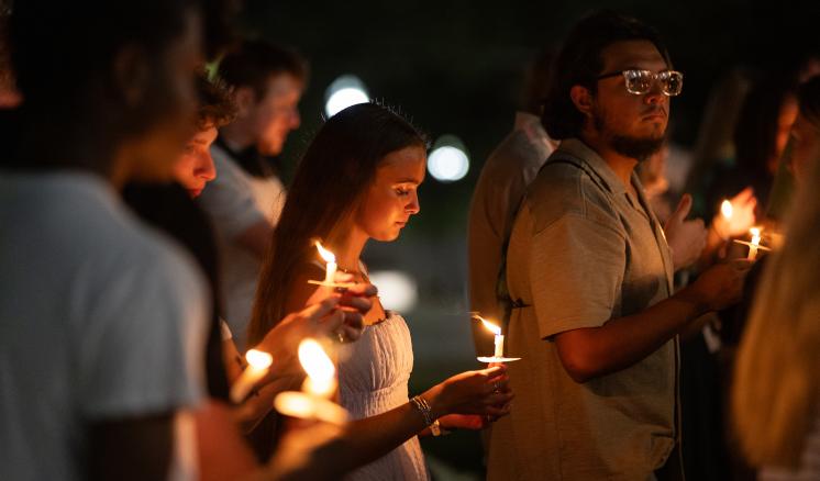 Photograph of a candlelight vigil held outdoors at night, showing a group of people holding lit candles with solemn expressions. Warm candlelight illuminates faces against a dark background, highlighting a sense of unity and remembrance.