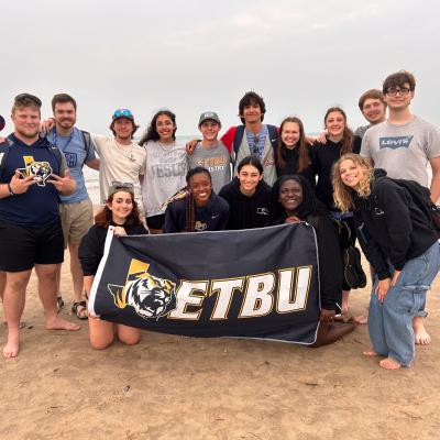 A group of individuals pose on the beach holding a flag with the ETBU logo on it