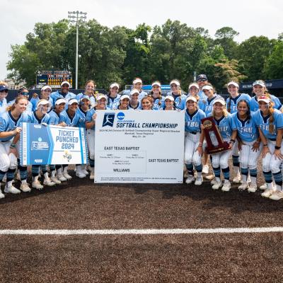 A large group photo of female softball players and coaches with a trophy in front of a score board on Taylor Field at East Texas Baptist University.