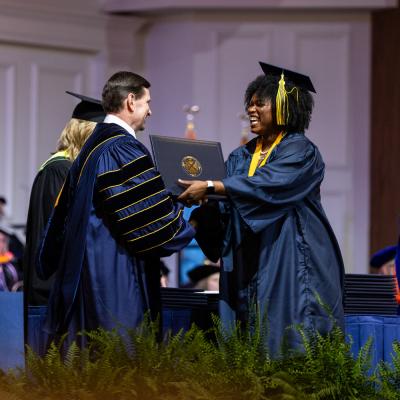 During a commencement ceremony, a woman in a navy cap and gown on the right receive her diploma from a man in navy academic regalia on the left.