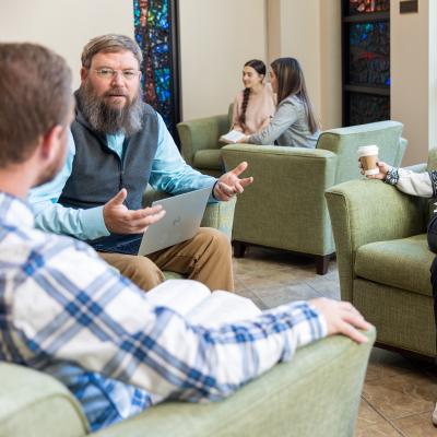 Faculty member sits in a common area conversing with students