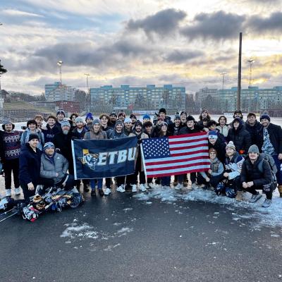 ETBU Hockey team stands in a large group outside in winter gear holding an ETBU branded flag and the American Flag